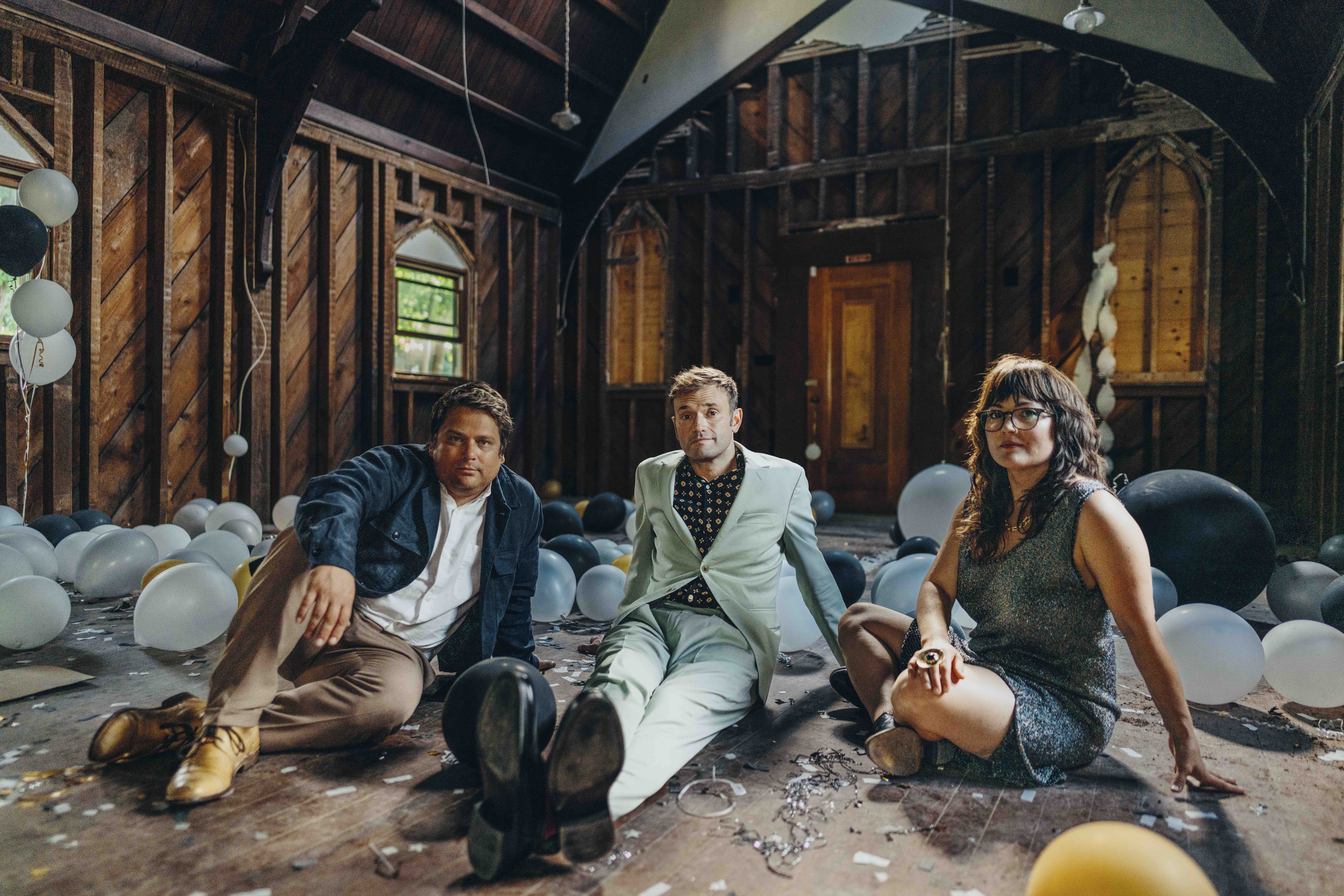 In an abandoned building with exposed wood framing, there are white and black balloons scattered on the floor. In the center, three people (those apart of Nickel Creek) sit next to one another.